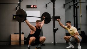 Muscular man lifting a barbell in a gym under his trainer's guidance, showcasing strength and focus.